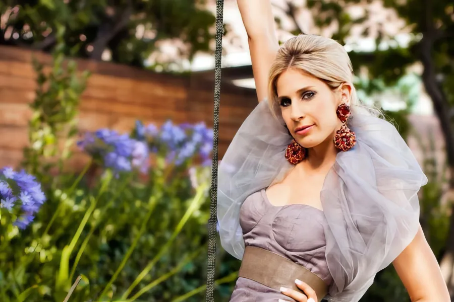 Soprano Hila Plitmann holds a rope outside while wearing a smoky lavendar gown with a billowing collar and large red gem earrings.