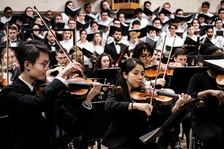 The first violin section of the University of Texas Symphony Orchestra plays, foregrounded by University of Texas choral singers.