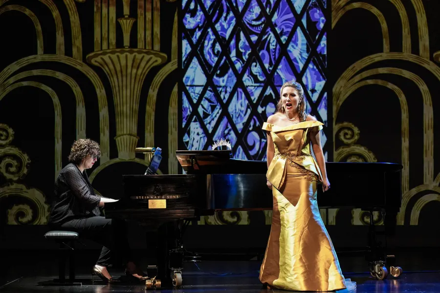Dr. Tamar Sanikidze plays piano while soprano Kara Covey sings wearing a gold gown. A stained glass window and pillars are projected behind them.