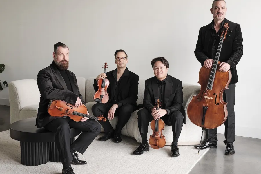 Members of the Miró Quartet wear black suits and gather around a white couch holding their instruments.