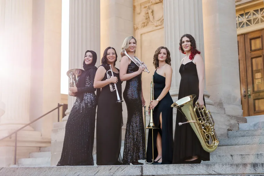 The members of Seraph Brass stand a grand stone staircase wearing black formal gowns, holding their instruments.