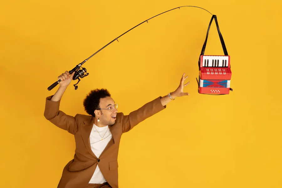 Accordion player Théo Ould reaches for a red accordion hanging from a fishing pole in front of a yellow background.