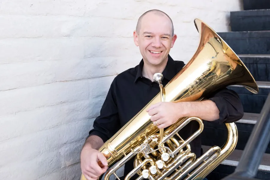Tuba player Scott Sutherland sits on a staircase holding his instrument in his lap.